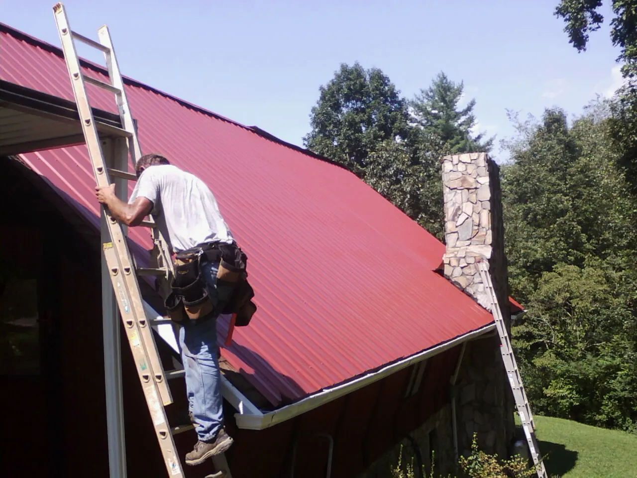 Roofer on a ladder beside a red metal roof and stone chimney — Soffit & Fascia Repair by Roofing Brothers in Oakdale